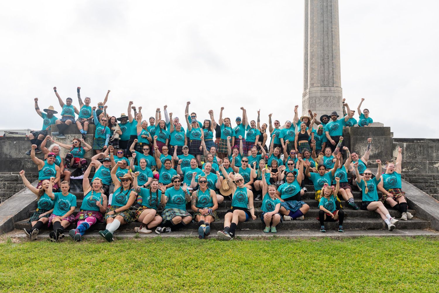 Large group of people in teal t-shirts celebrating on monument steps outdoors.