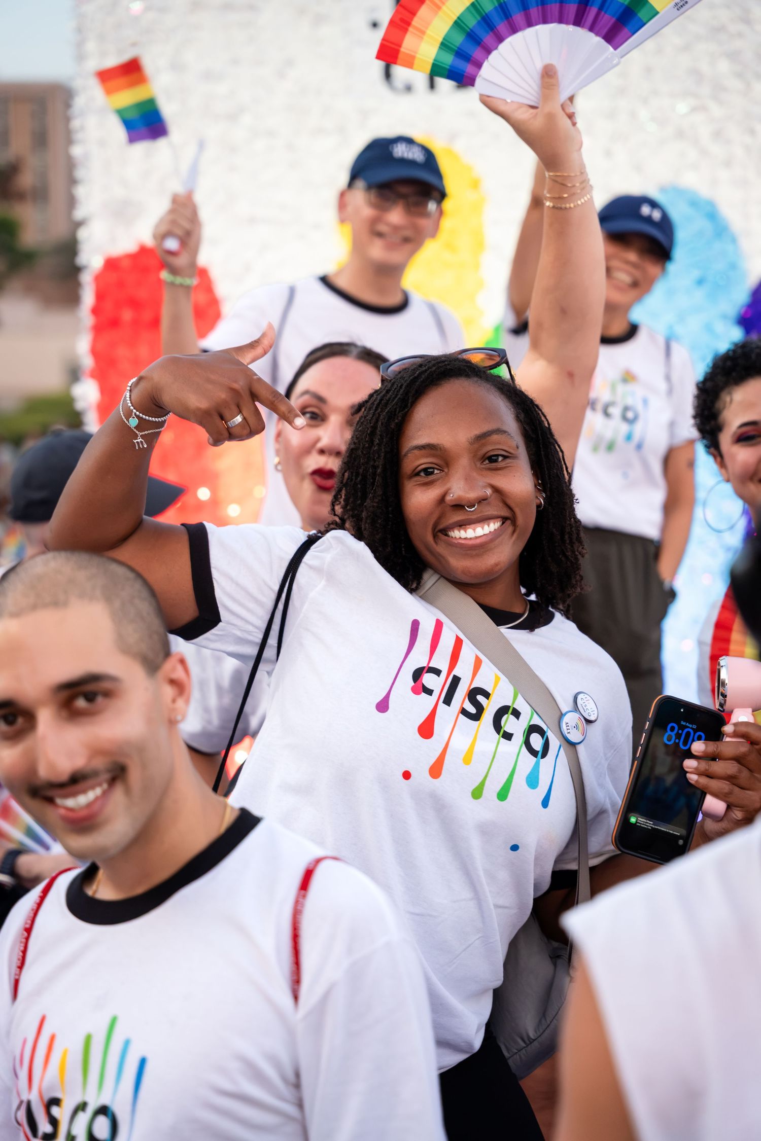 Diverse group of smiling people in Cisco rainbow logo shirts celebrating Pride parade, holding rainbow flags and fans.