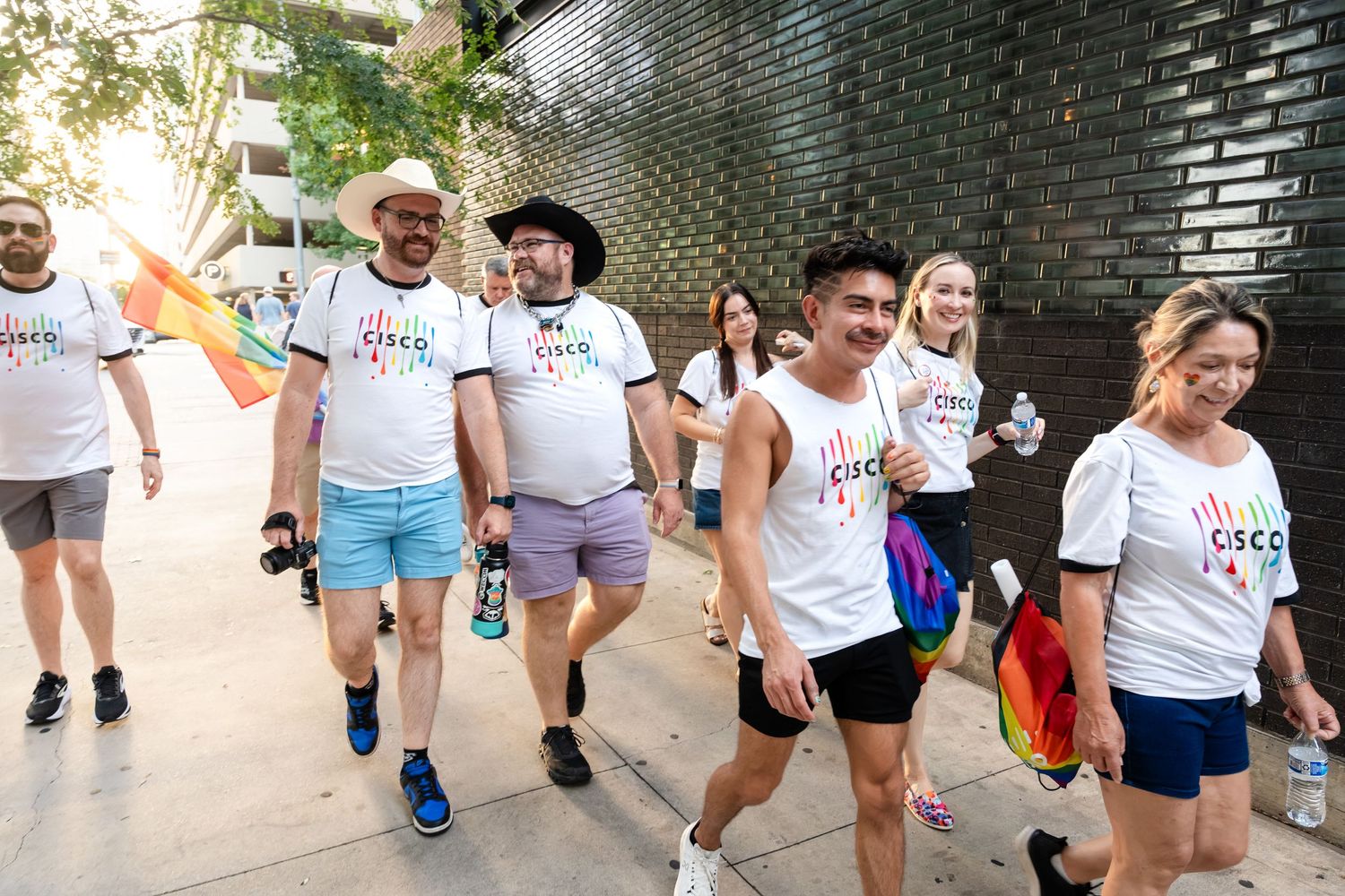 Group of Cisco employees wearing branded white t-shirts walking together in a Pride parade, carrying rainbow flags.