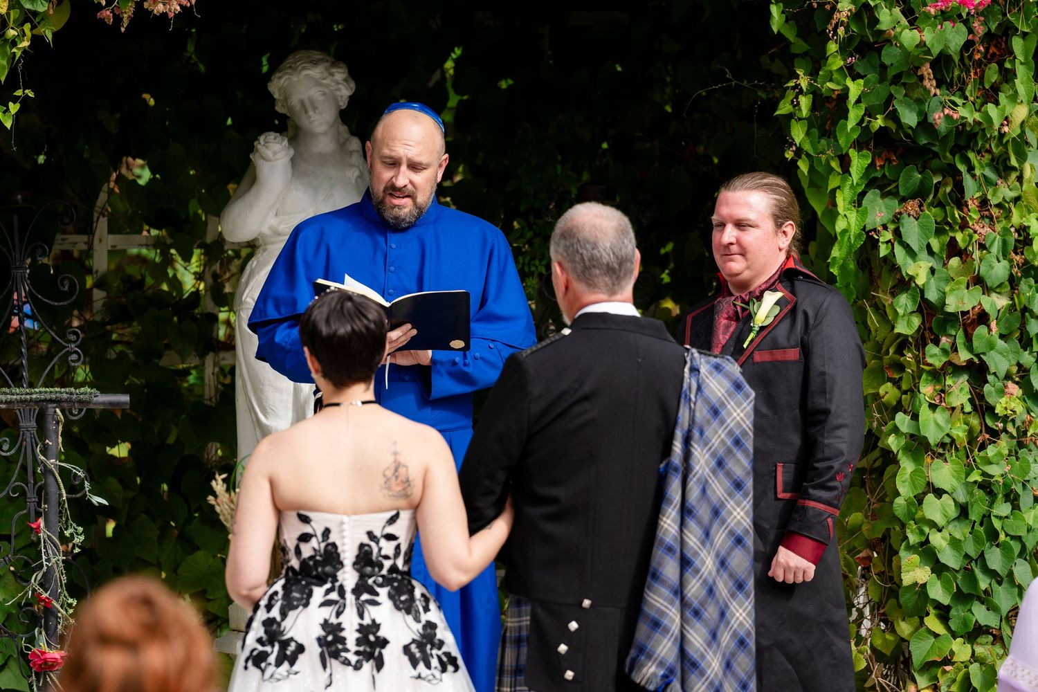 Outdoor garden wedding ceremony with officiant in blue and couple standing among ivy-covered walls and stone statues.