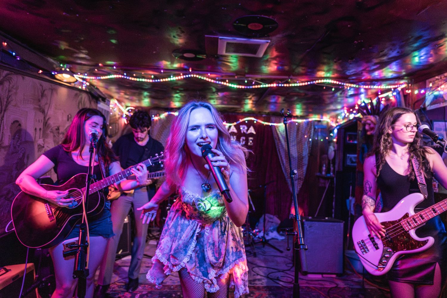 Musicians perform on stage in a dimly lit venue with colorful purple and blue lighting creating an atmospheric ambiance.