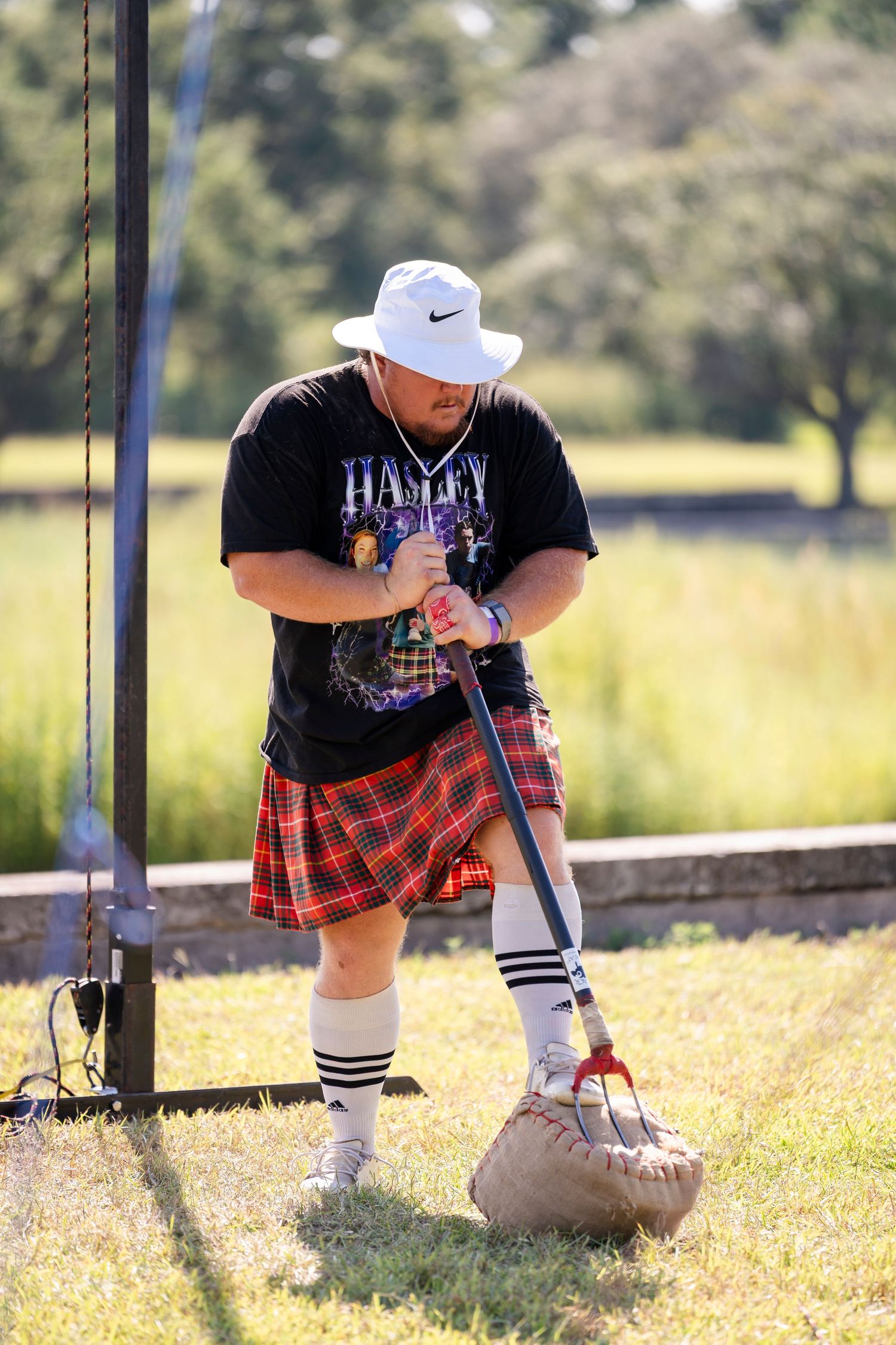 A person in a black t-shirt and red plaid skirt sits on a wooden fence in a sunny outdoor field.