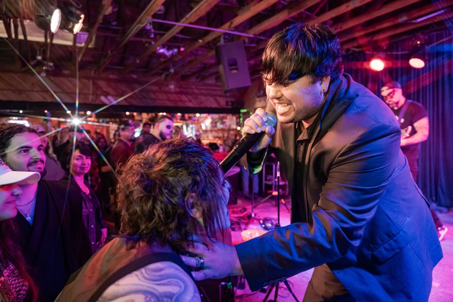 A performer in a blue jacket sings passionately into a microphone at an intimate concert venue with purple lighting.