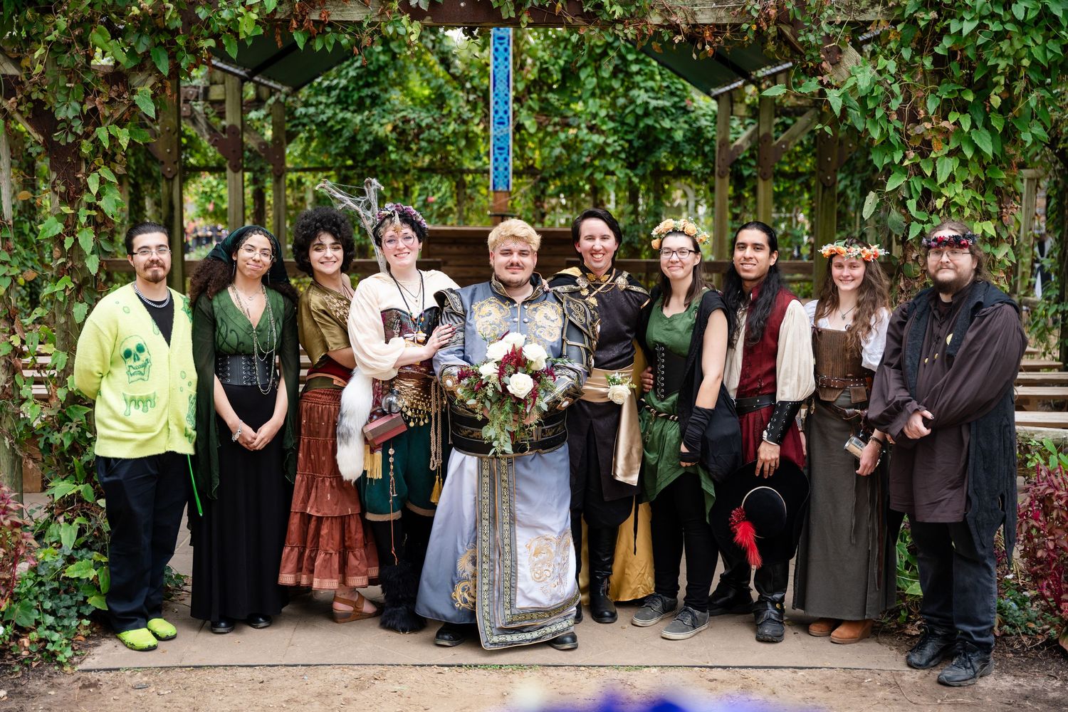 A group of people stand in a line outdoors against a green foliage backdrop wearing various casual attire.