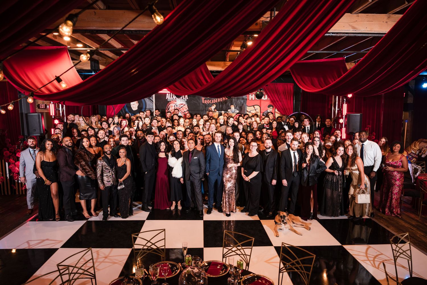 Large formal event with guests in elegant attire gathered on a black and white checkered dance floor under red draping.