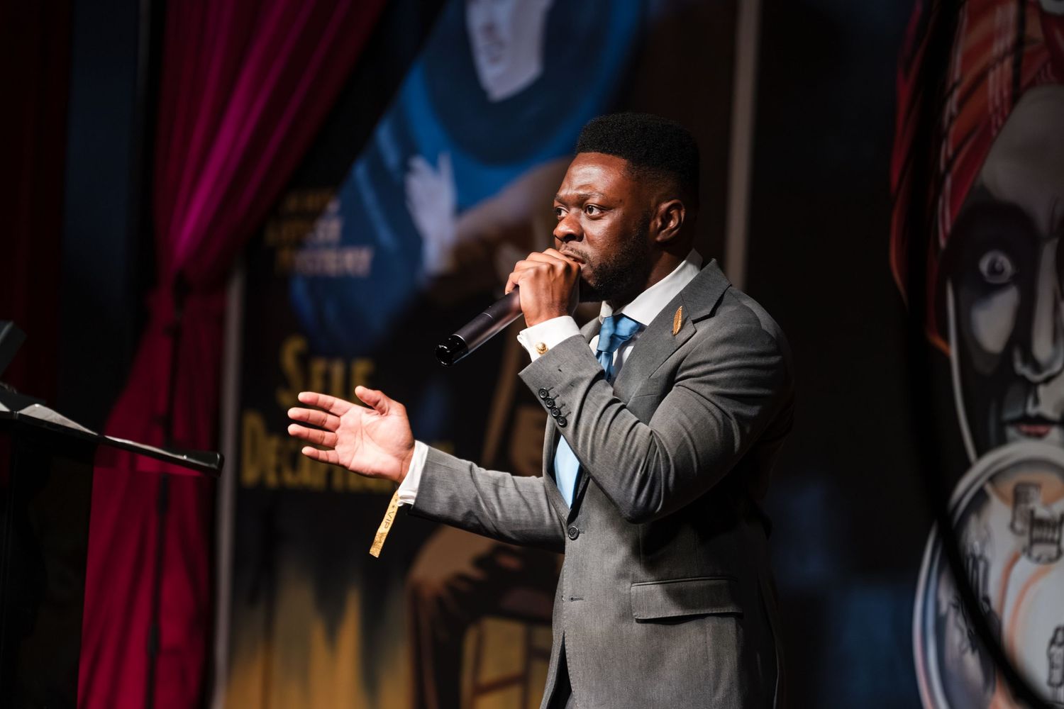 A performer in a dark suit sings passionately on stage with dramatic lighting in the background.
