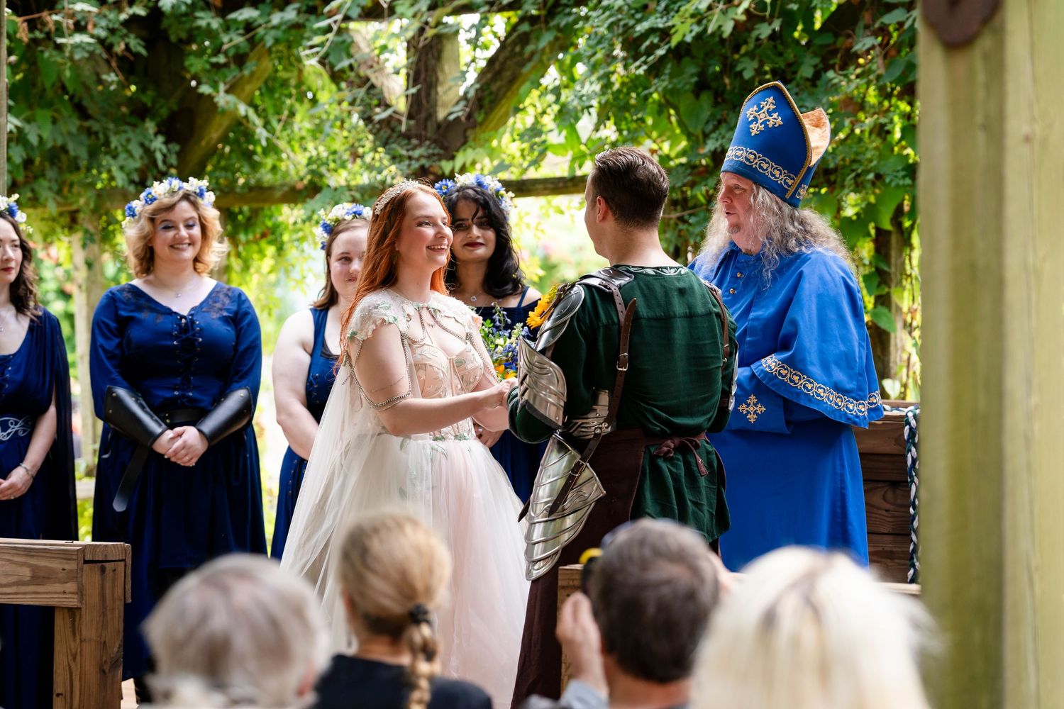 A couple exchanges vows during an outdoor wedding ceremony under a wooden pergola with guests in blue attire watching.