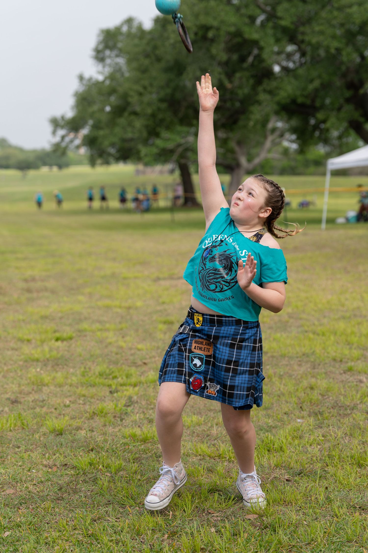 Young girl in tartan kilt and teal shirt throwing a ball at an outdoor Scottish Highland Games competition.