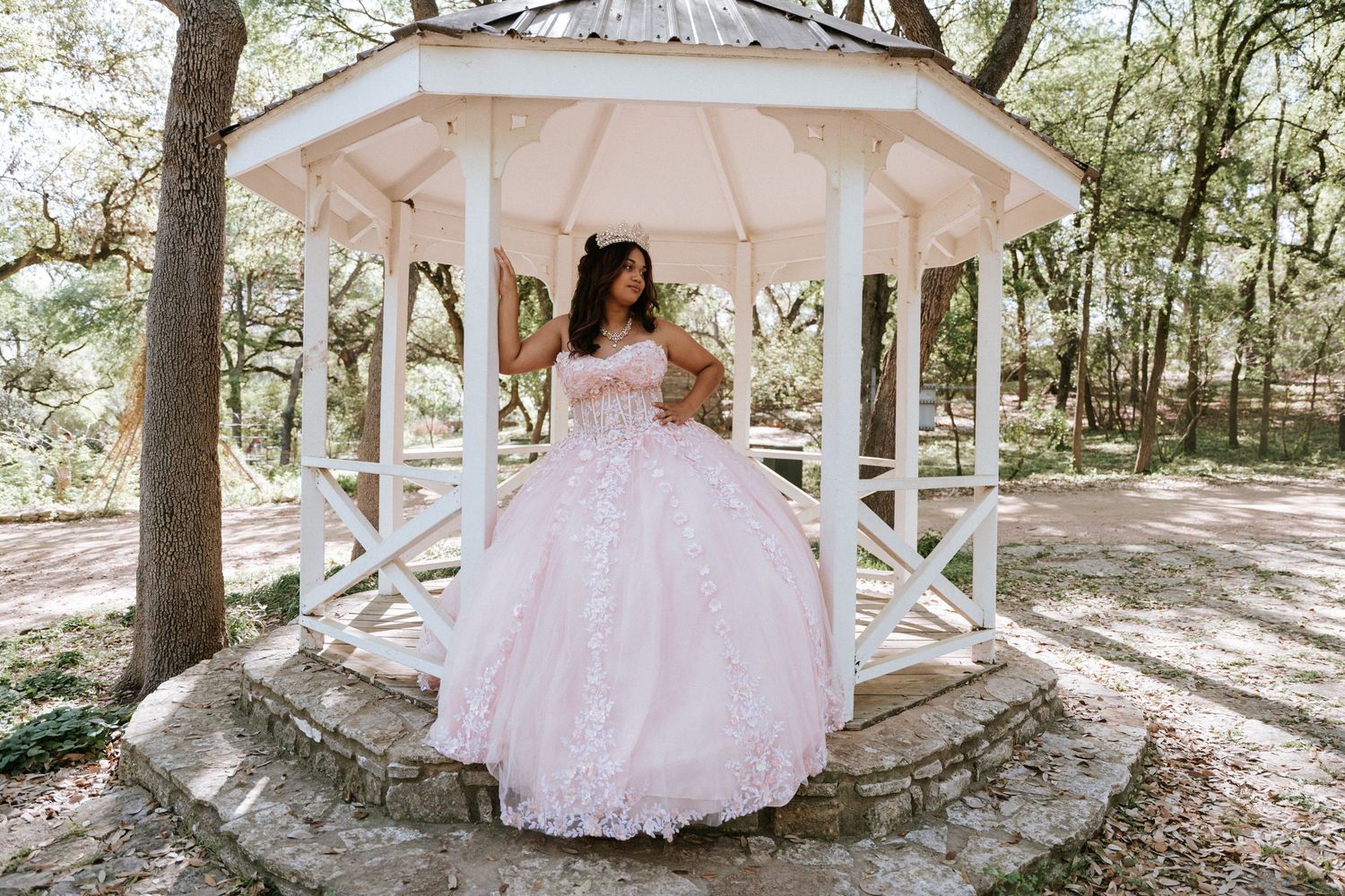 Young woman in pink ball gown poses gracefully under white gazebo surrounded by trees in a park setting.
