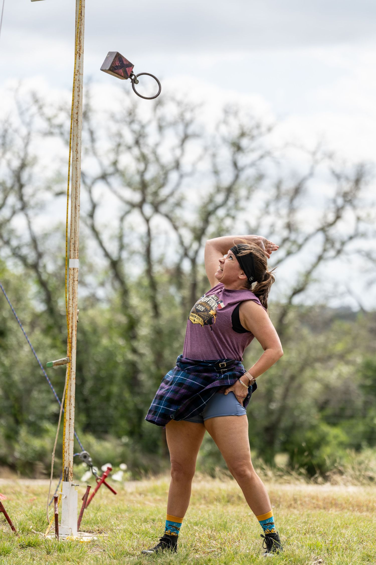 Woman in plaid skirt and purple top looks up at pole during outdoor Highland Games competition.
