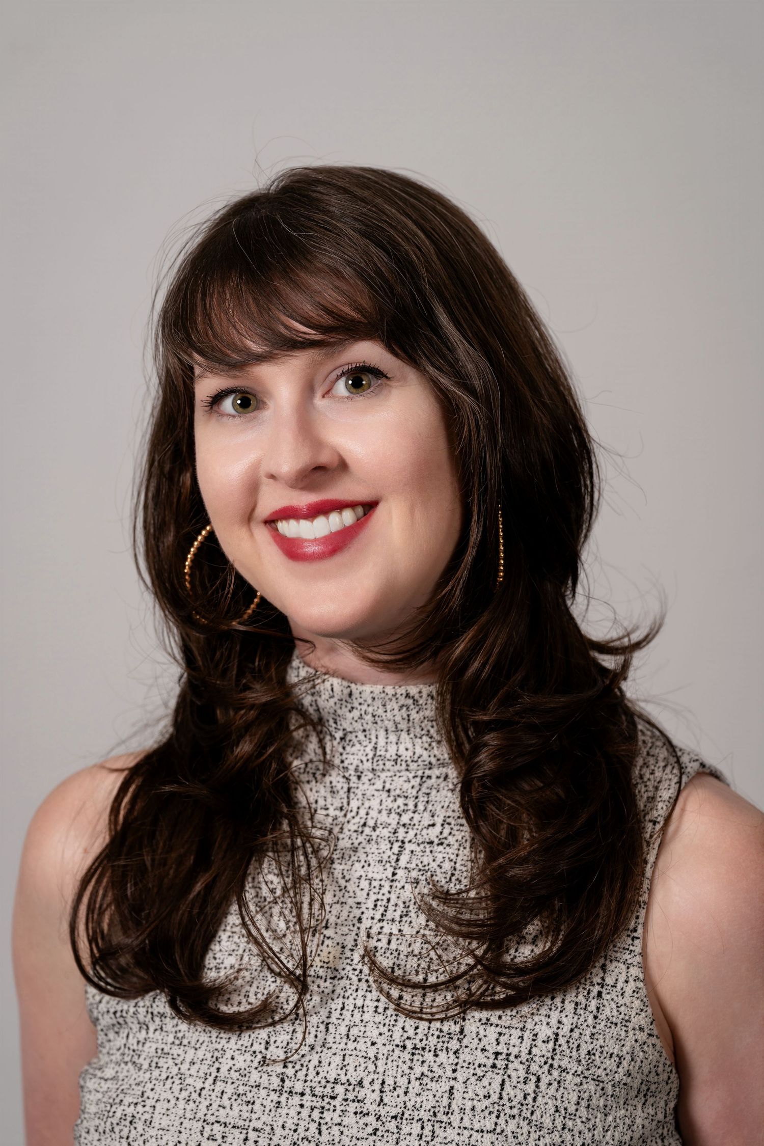 Smiling woman with dark hair, red lips, gold hoop earrings, wearing a sleeveless gray tweed top against a neutral background.