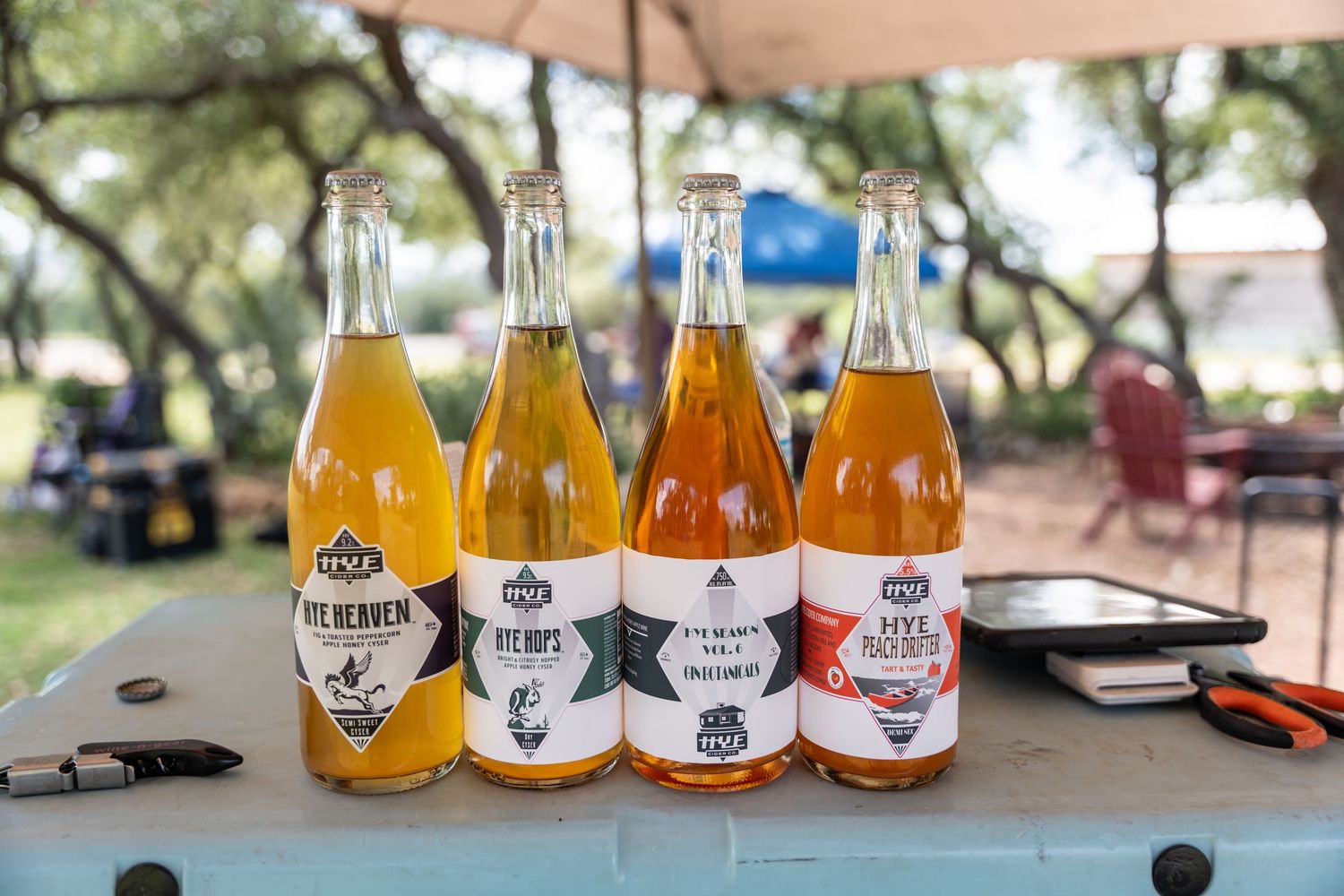 Four craft cider bottles with colorful labels displayed on a table outdoors at a festival event.