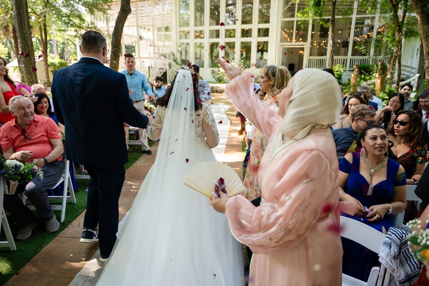 Outdoor wedding ceremony with bride in white gown and veil, groom in suit, guests seated, and women in pink hijabs celebrating.