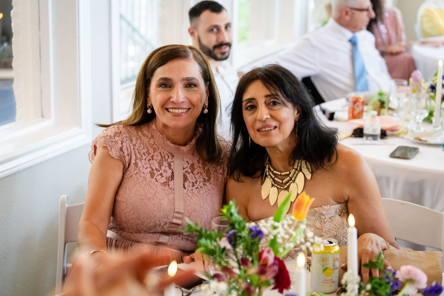Two women smiling together at an elegant wedding reception dinner table with floral centerpieces.