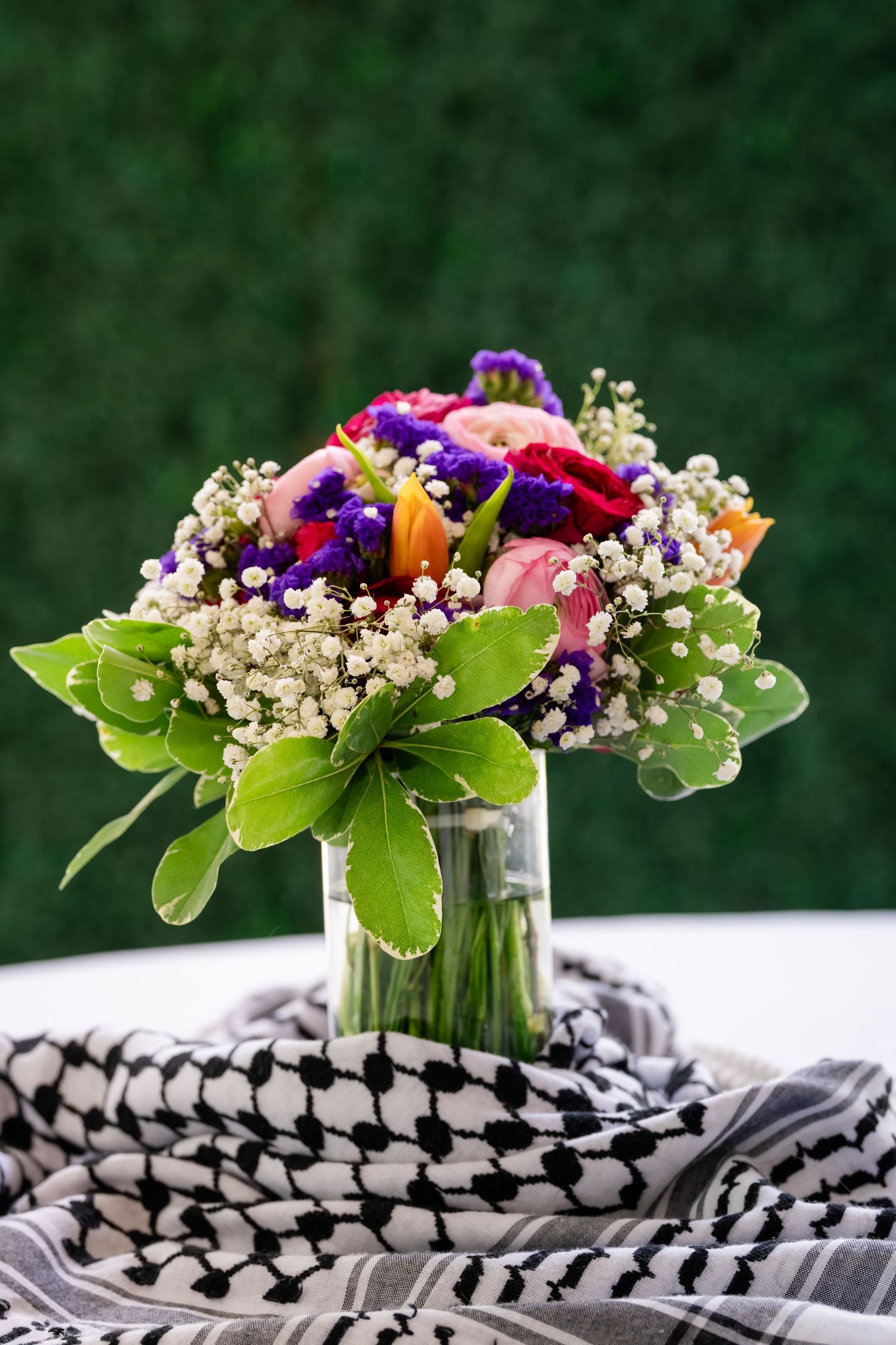 Colorful bridal bouquet with pink roses, purple flowers, and baby's breath in a glass vase on black and white fabric.