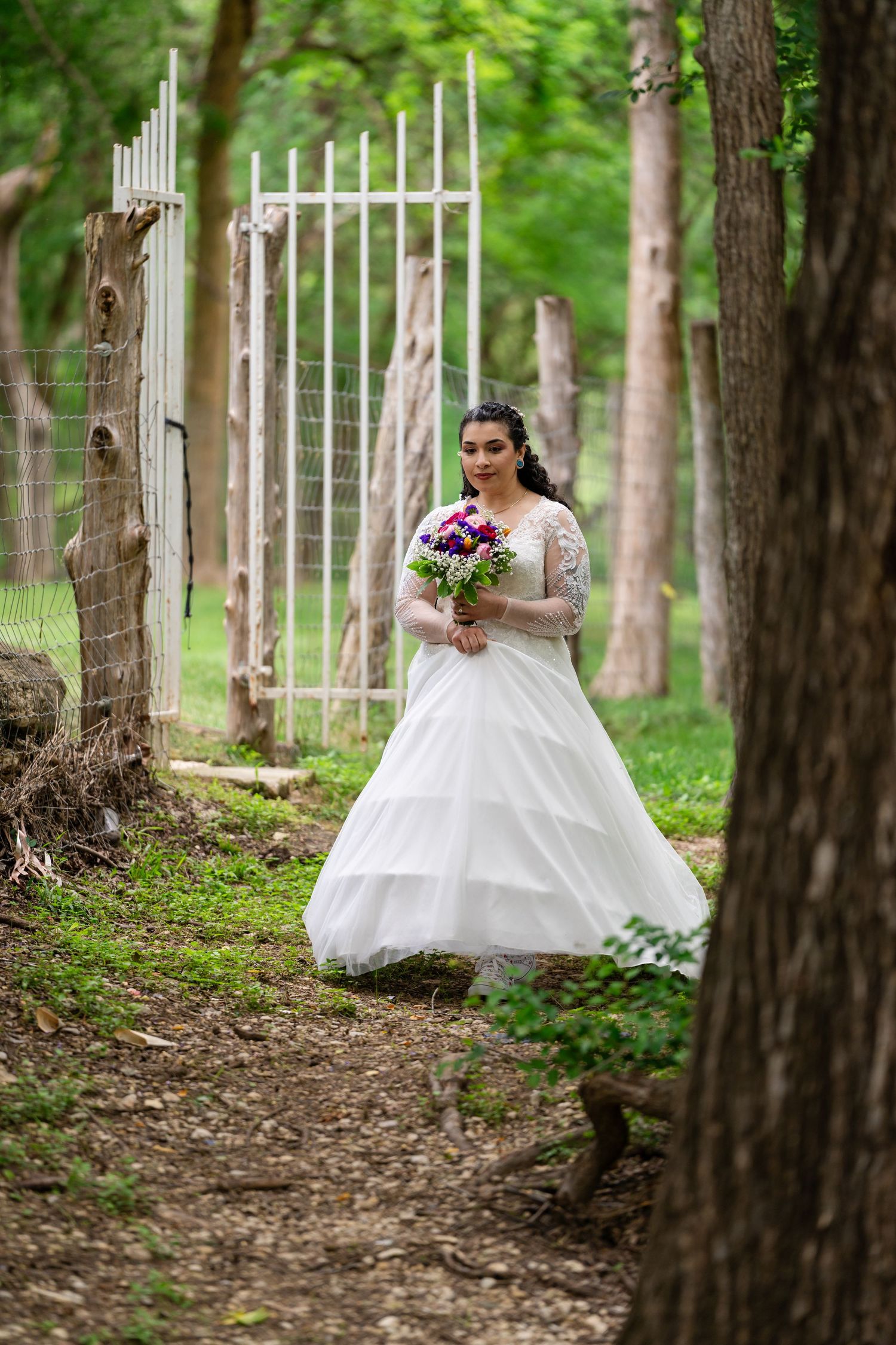 Bride in white lace gown holding purple bouquet walks along wooded path near white gate.