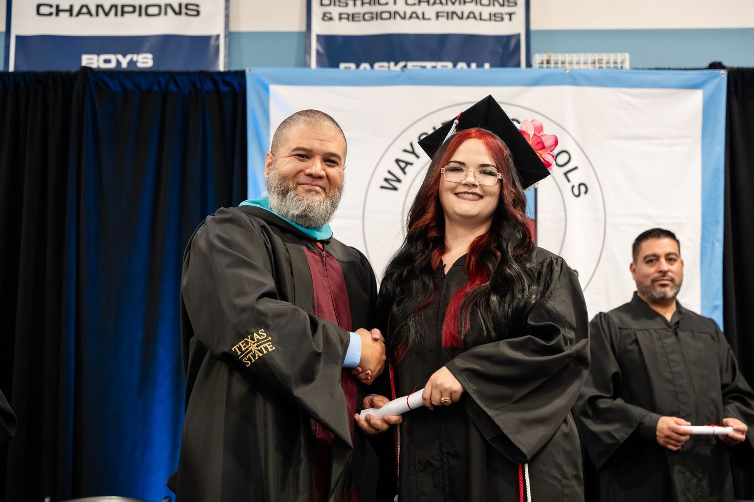Two graduates in black robes shake hands on stage during a school graduation ceremony with banners in the background.