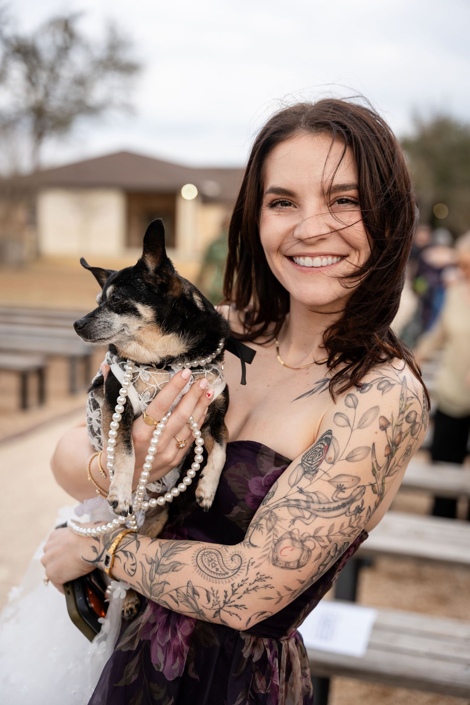 Smiling woman with tattoos holding a small dog wearing pearl necklaces outdoors.