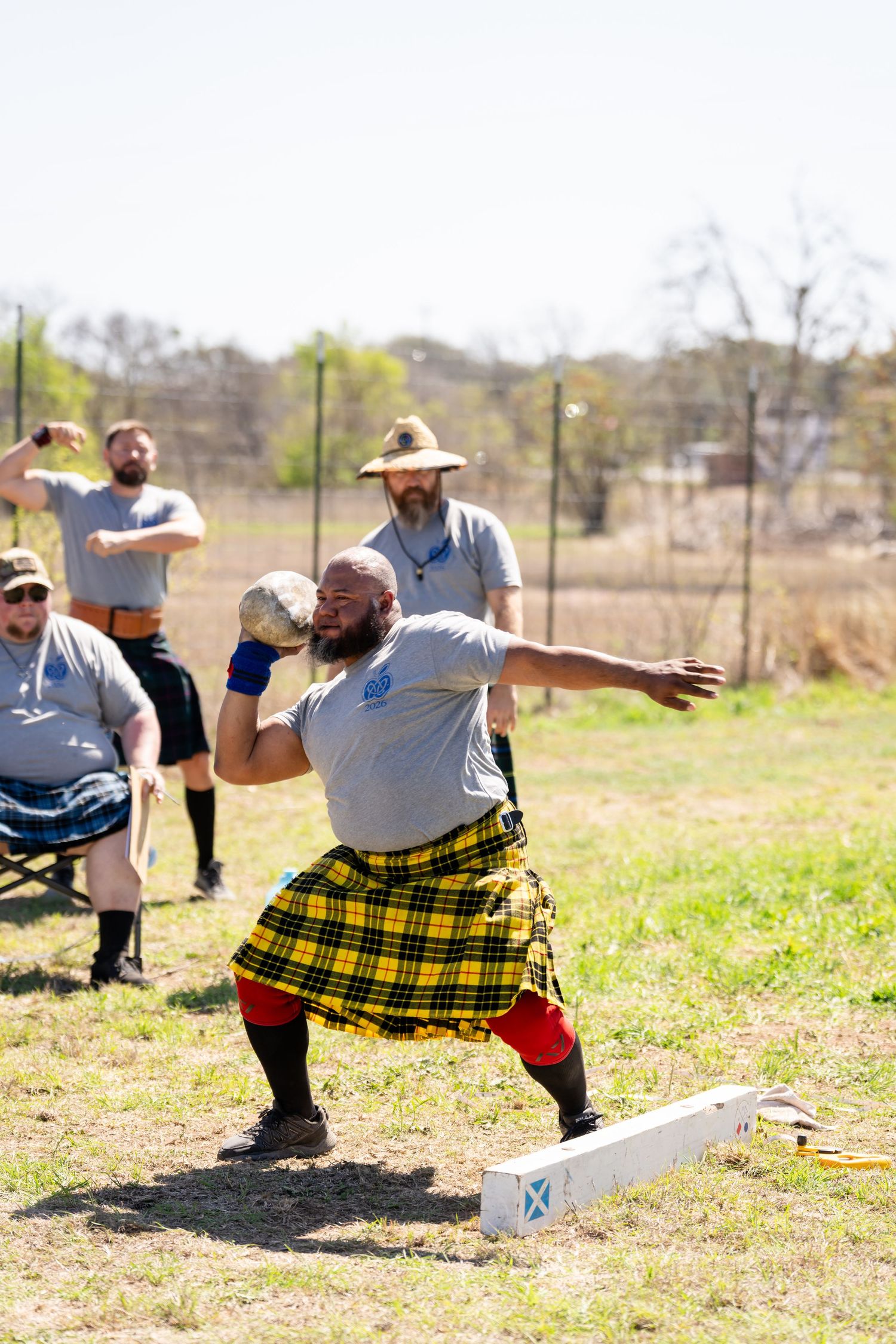 Man in yellow tartan kilt throws shot put at Scottish Highland Games competition outdoors.