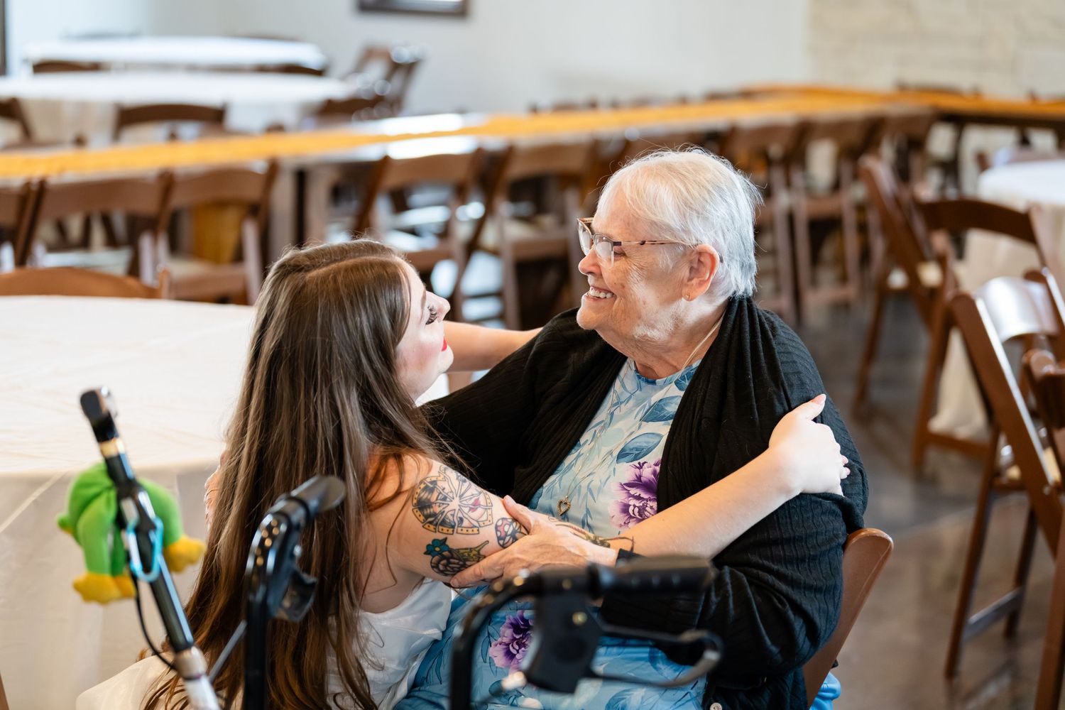 Elderly woman and young woman with tattoos sharing a warm embrace and smile in a banquet hall.