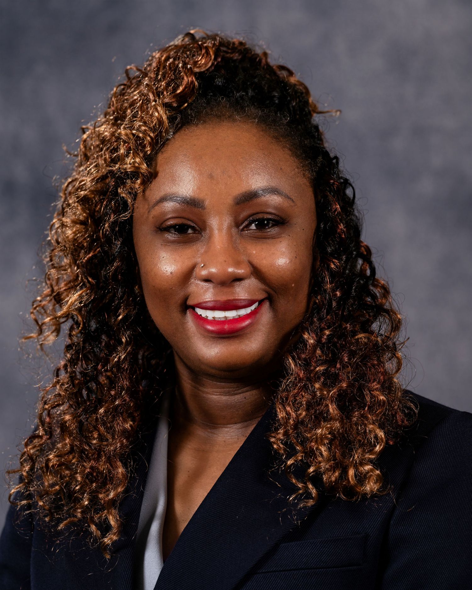Professional headshot of a smiling woman with curly auburn hair wearing a dark blazer against a gray background.