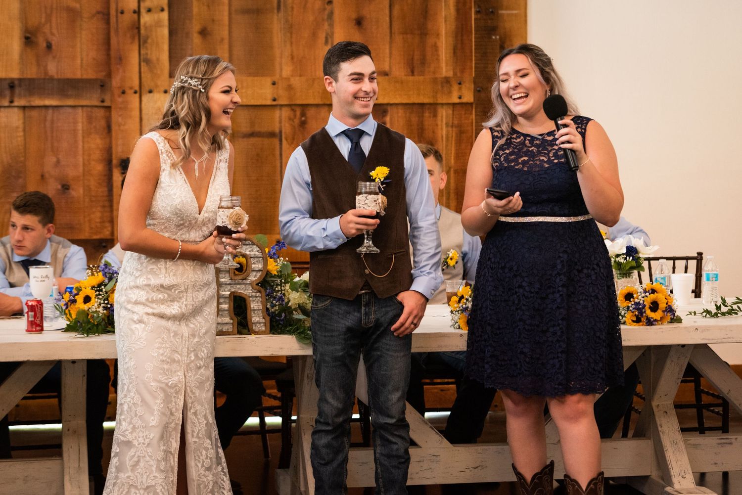 Three wedding party members share laughter while holding drinks at reception with rustic barn backdrop.