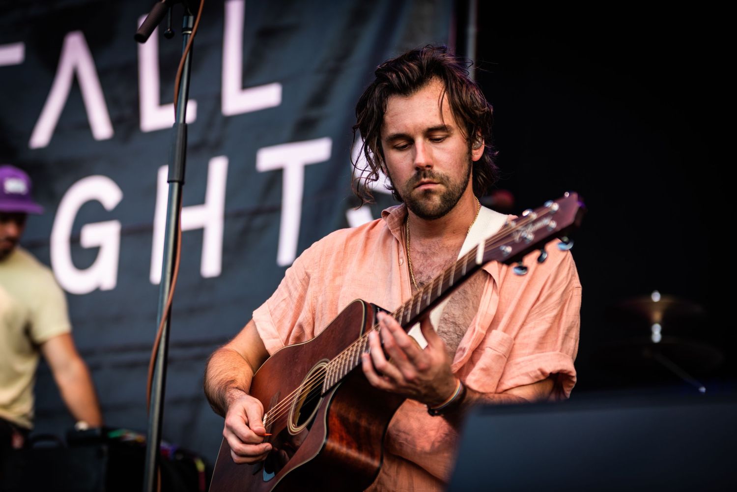 Musician performs on acoustic guitar wearing a pink button-down shirt during outdoor concert.