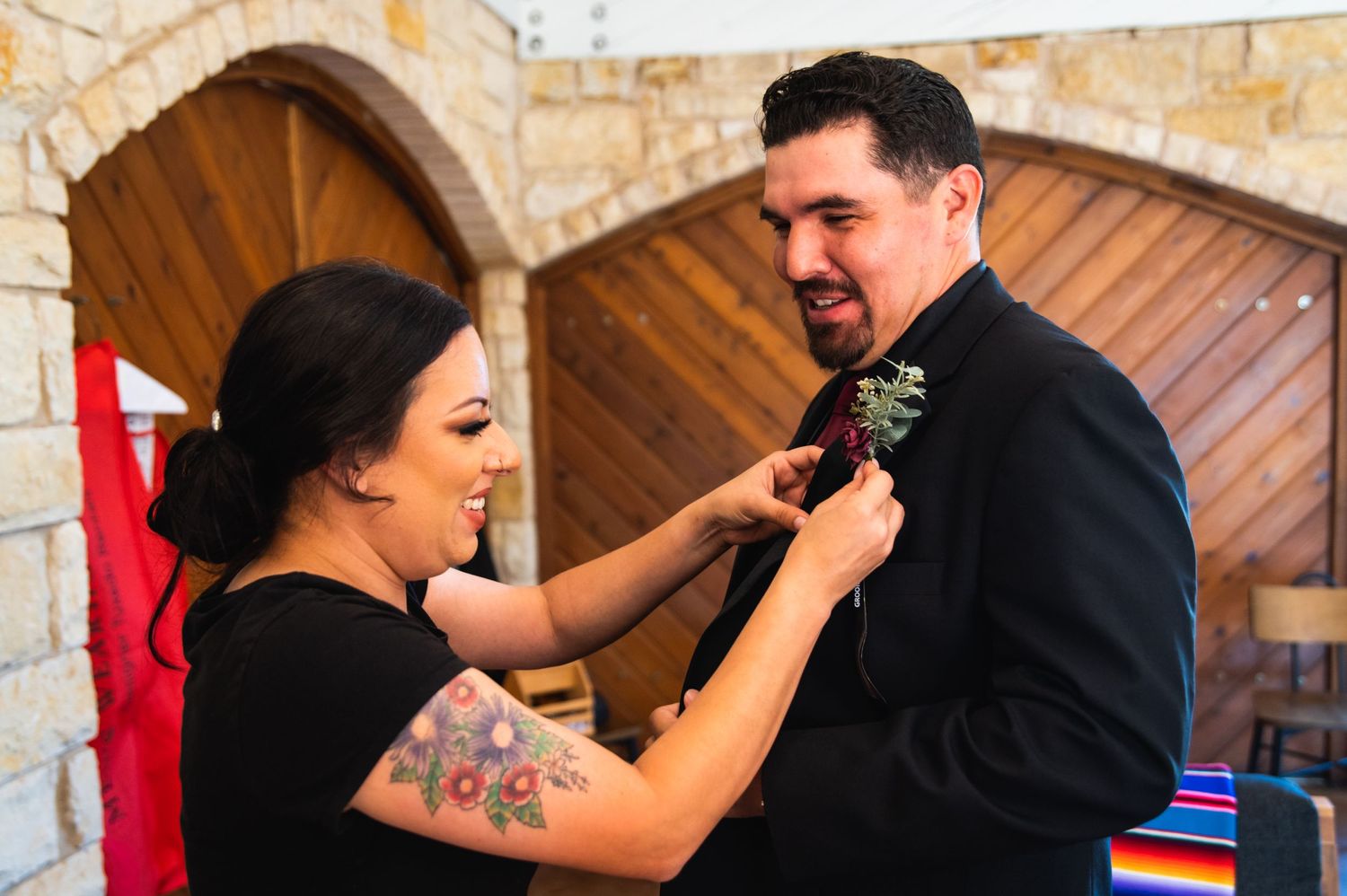 A person with tattoos pins a boutonniere on someone wearing a black suit.