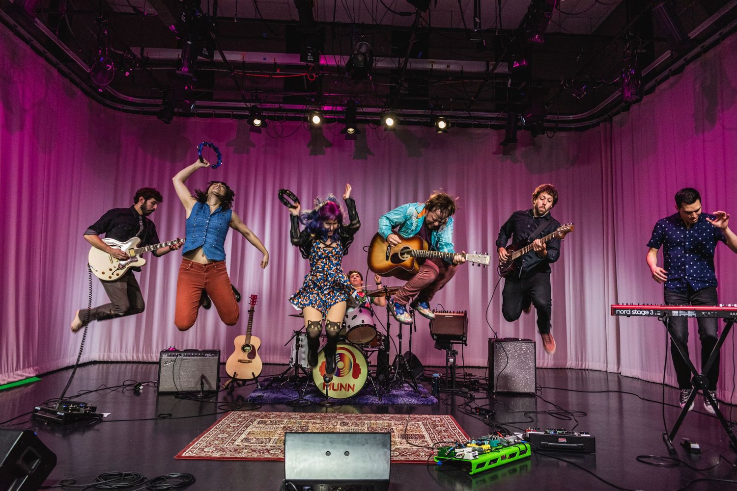 Live band performs on stage with instruments and equipment against pink curtain background.