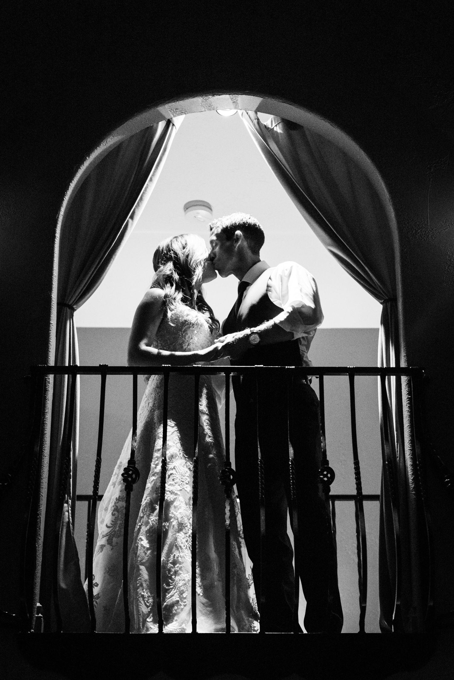 A romantic silhouette of a couple sharing a moment on a balcony through an arched window.