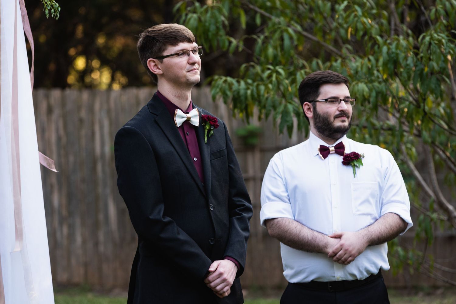 Two people in formal attire stand outdoors during a ceremony, wearing matching burgundy bow ties.