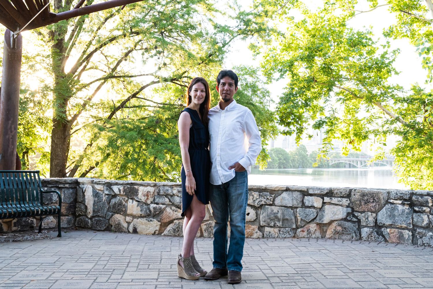 A sunny engagement photo taken at a stone pavilion with lush green trees.