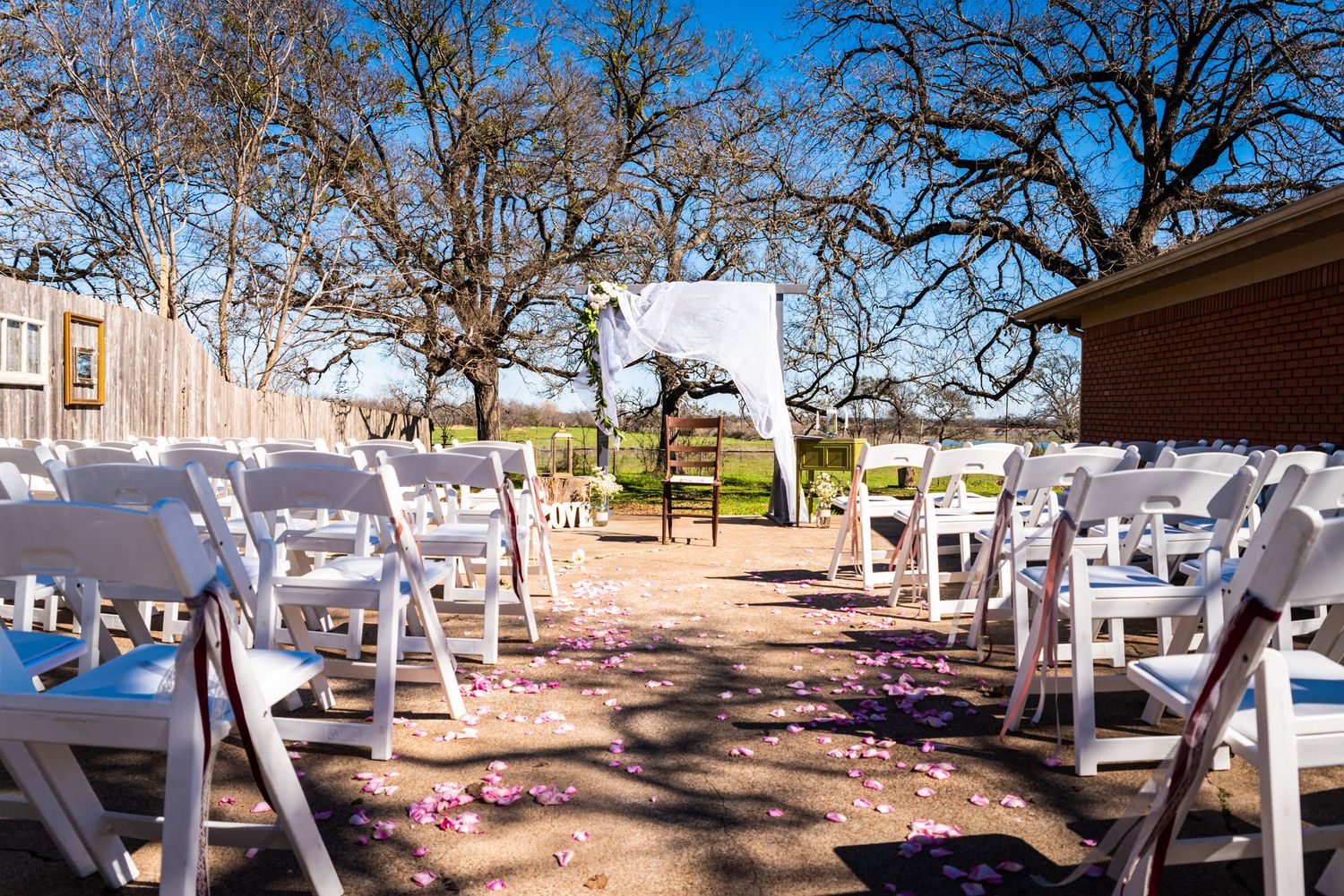 White chairs line an outdoor wedding ceremony aisle with rose petals beneath bare winter trees.