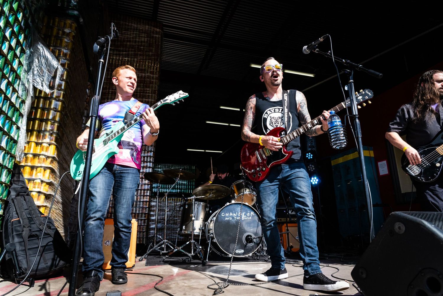 A band performs on an outdoor stage with guitars and drums during a daytime concert.