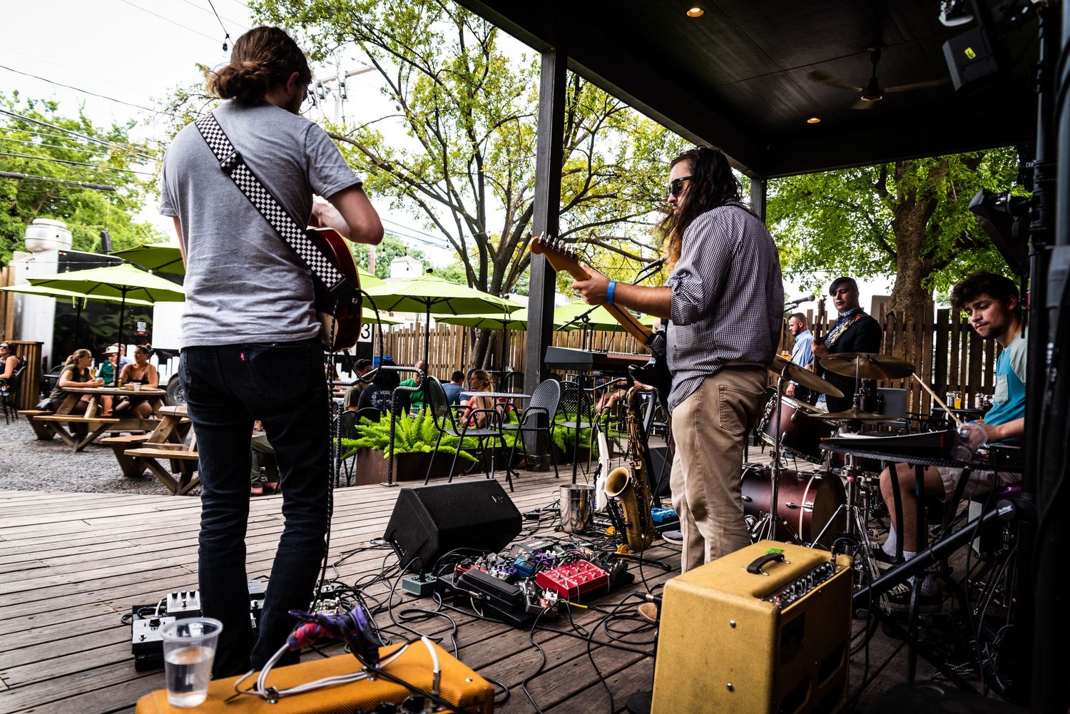 Musicians perform on outdoor wooden deck stage with equipment and trees in background.