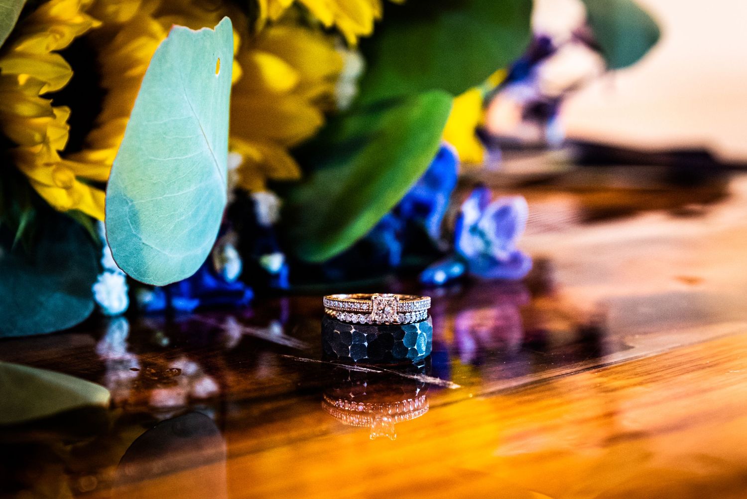 Wedding rings on a reflective surface next to colorful flowers.