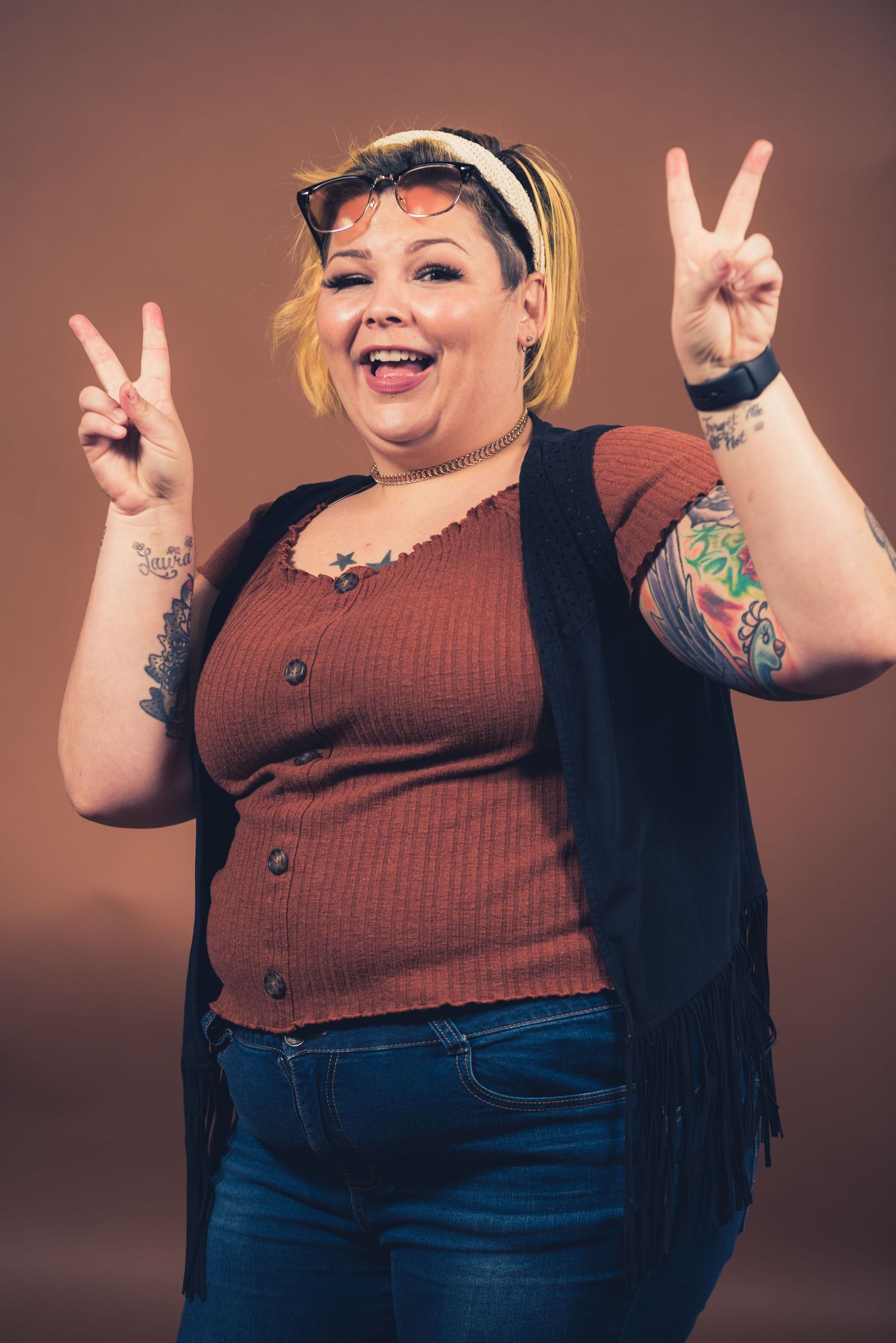 Happy person in brown top and black vest making peace signs against brown background.