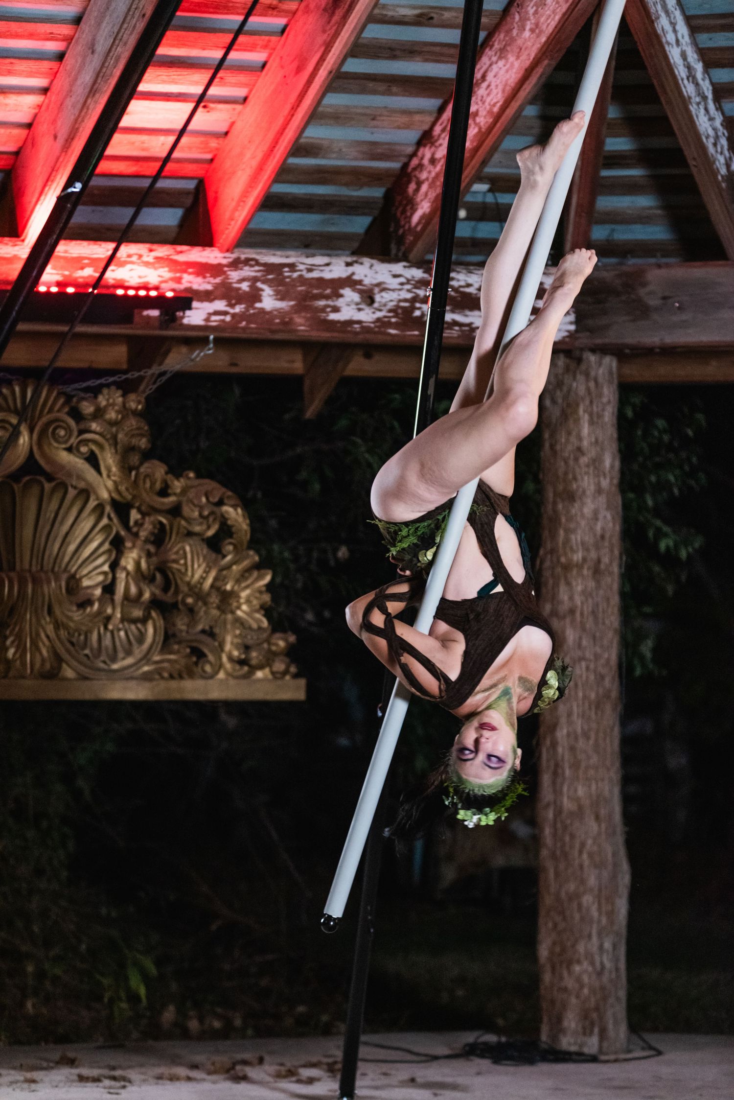 Performer executes inverted aerial pole move under red-lit wooden rafters.
