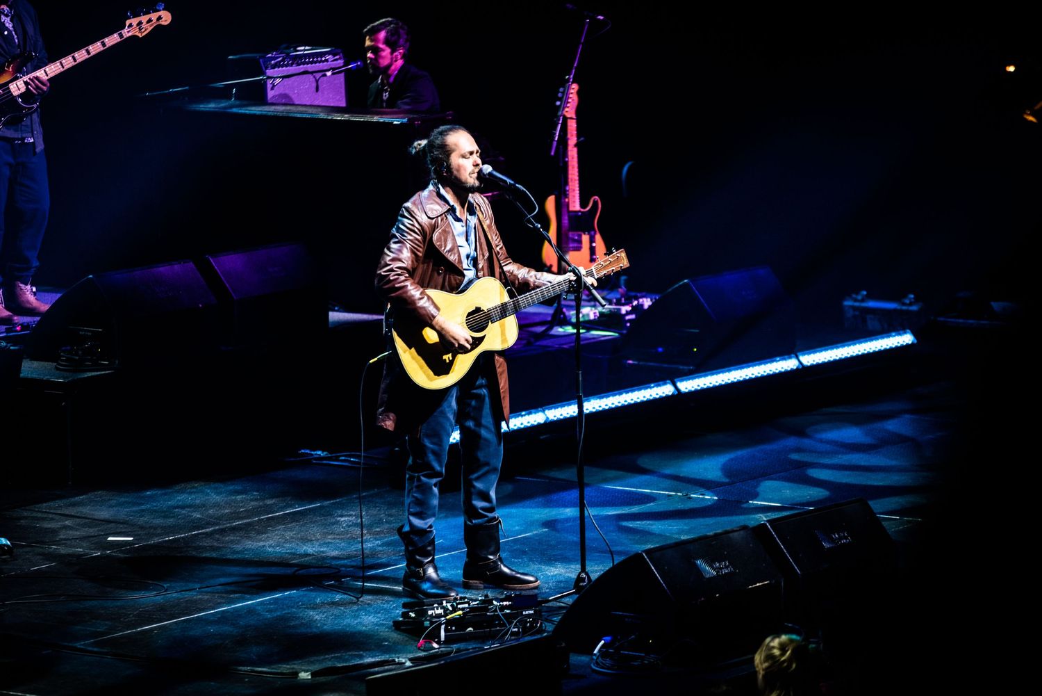 Musician plays acoustic guitar on darkened stage with instrumental setup visible behind.