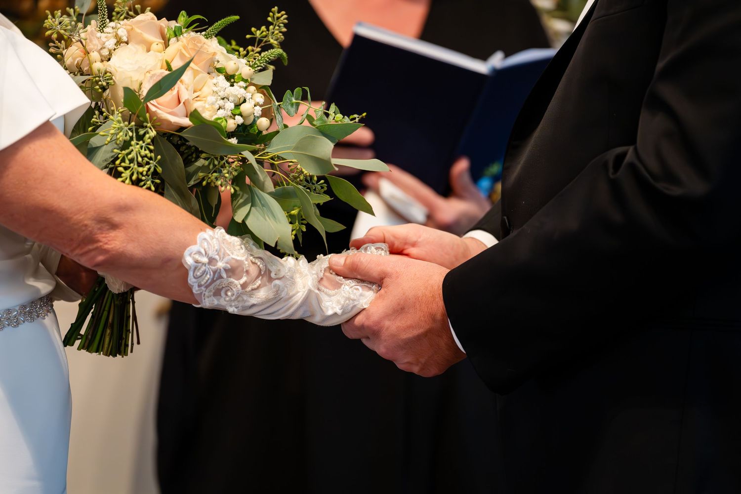 Close-up of hands during wedding ring exchange with lace sleeve detail and floral bouquet.