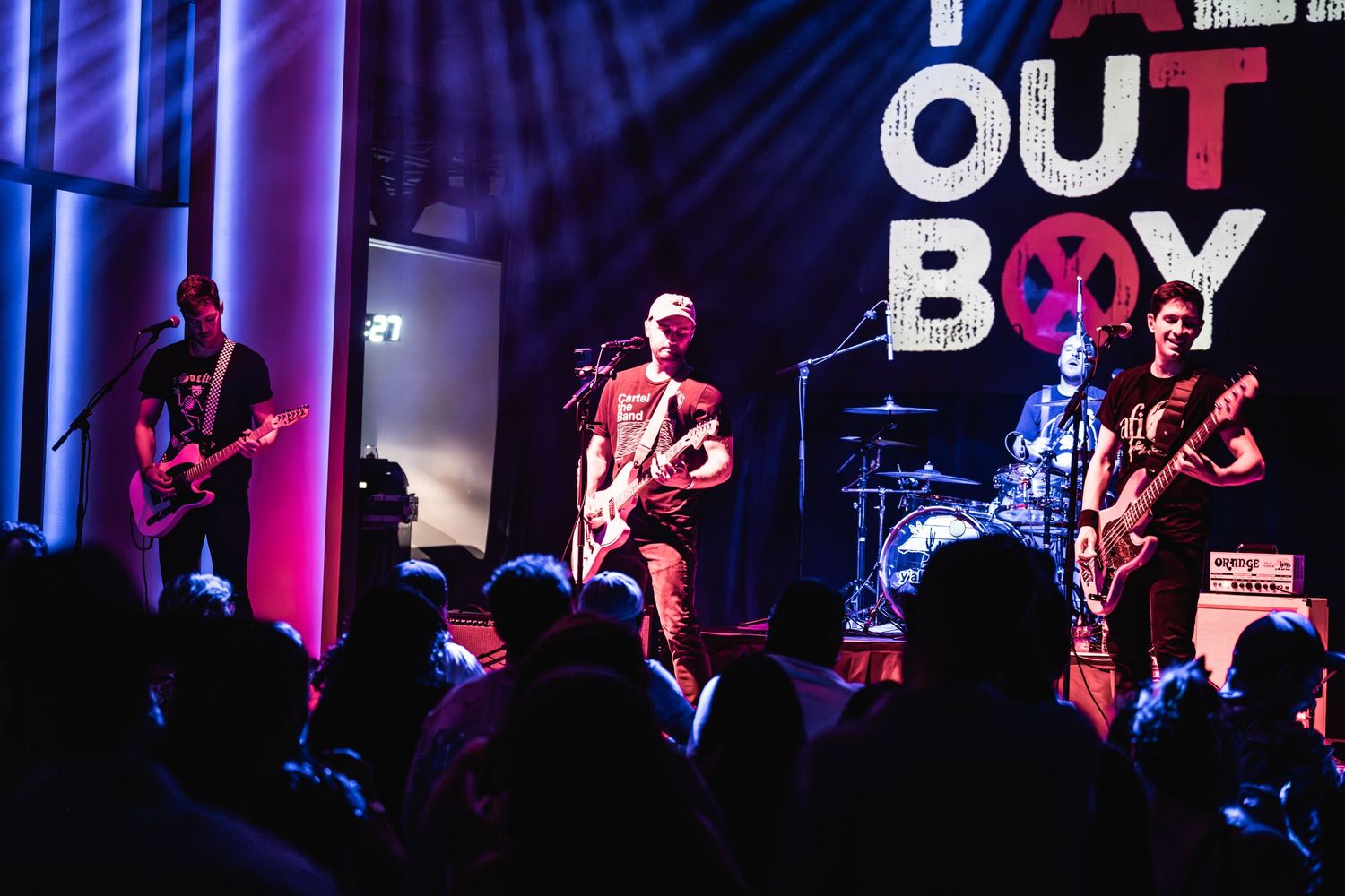 Rock band performs energetically on stage with purple lighting and silhouetted crowd at concert venue.