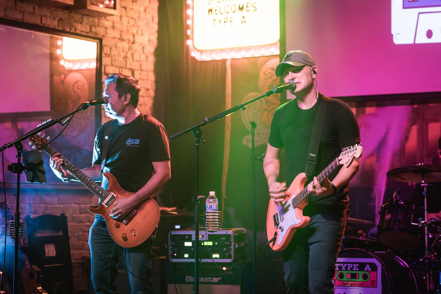 Two guitarists perform on stage with microphones under colorful venue lighting.