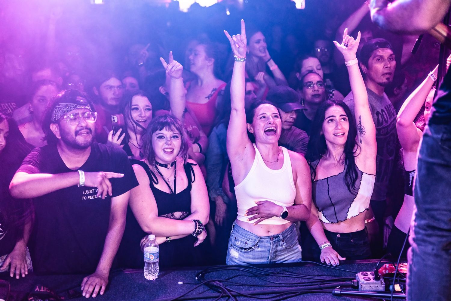 Concert crowd cheering with raised hands under purple stage lights at a music venue.