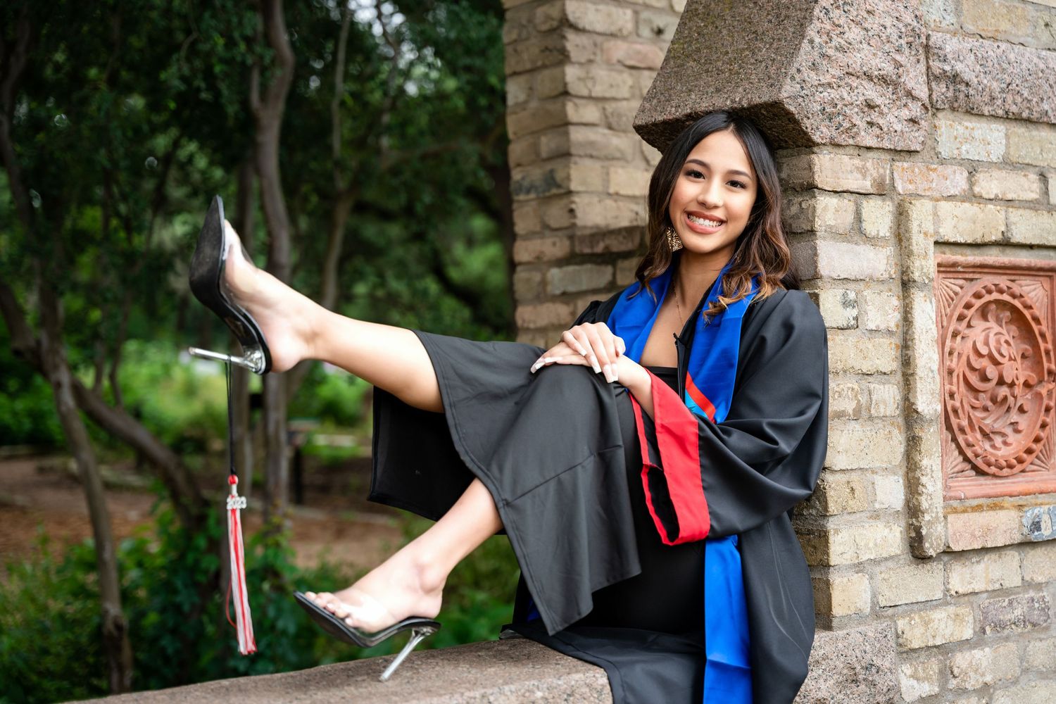 Graduate in cap and gown shows off diploma while posing on stone ledge outdoors.