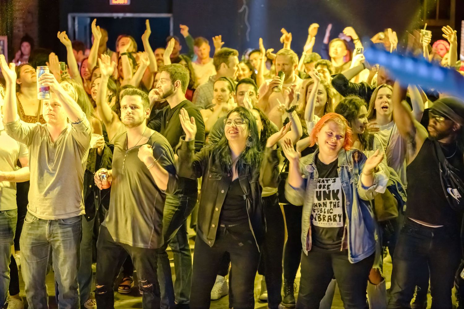 Energetic crowd with raised arms cheering at concert under yellow lighting.