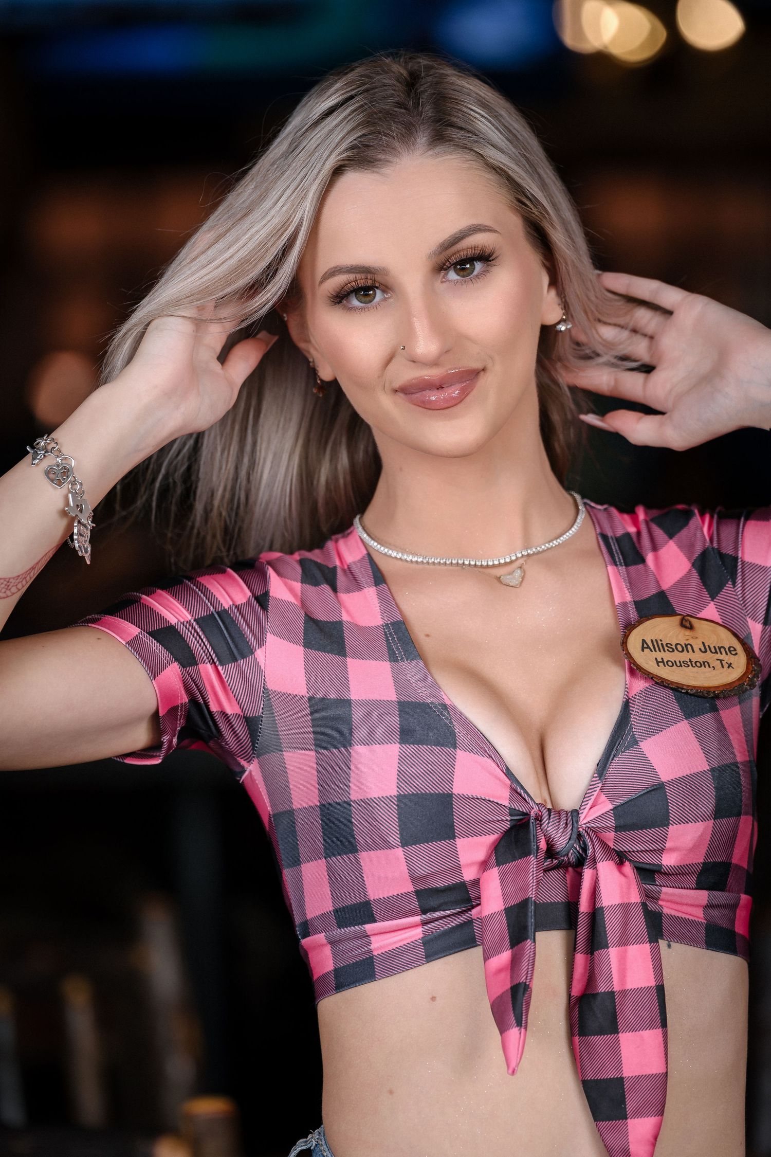 Woman in pink plaid crop top poses with hands in hair and necklace against dark background.