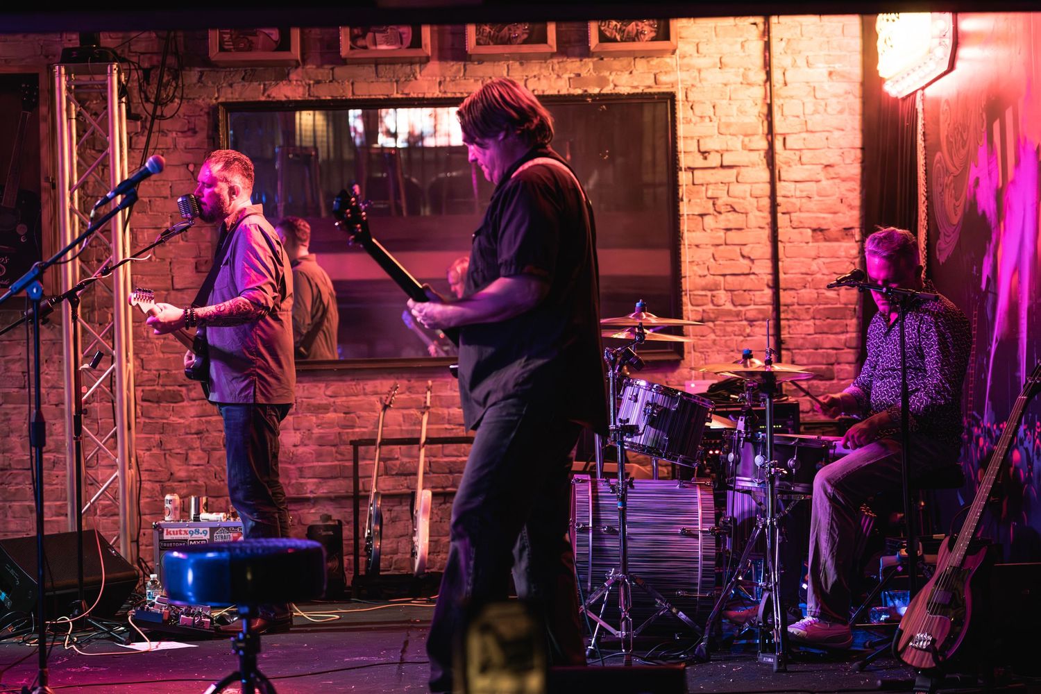 Three-piece band performs on brick-walled stage under moody lighting.