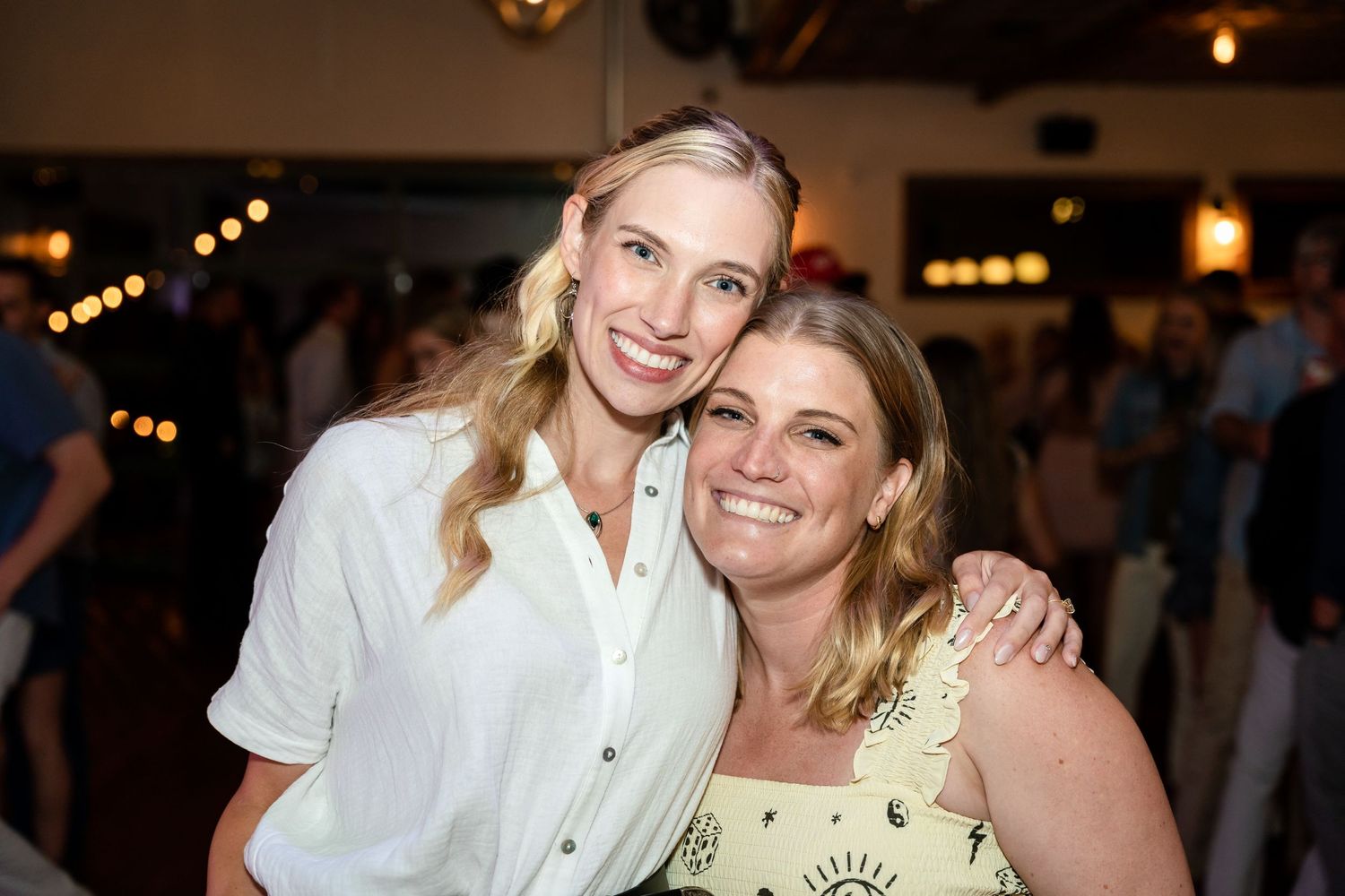 Two friends sharing a warm moment at an indoor social gathering with ambient lighting.