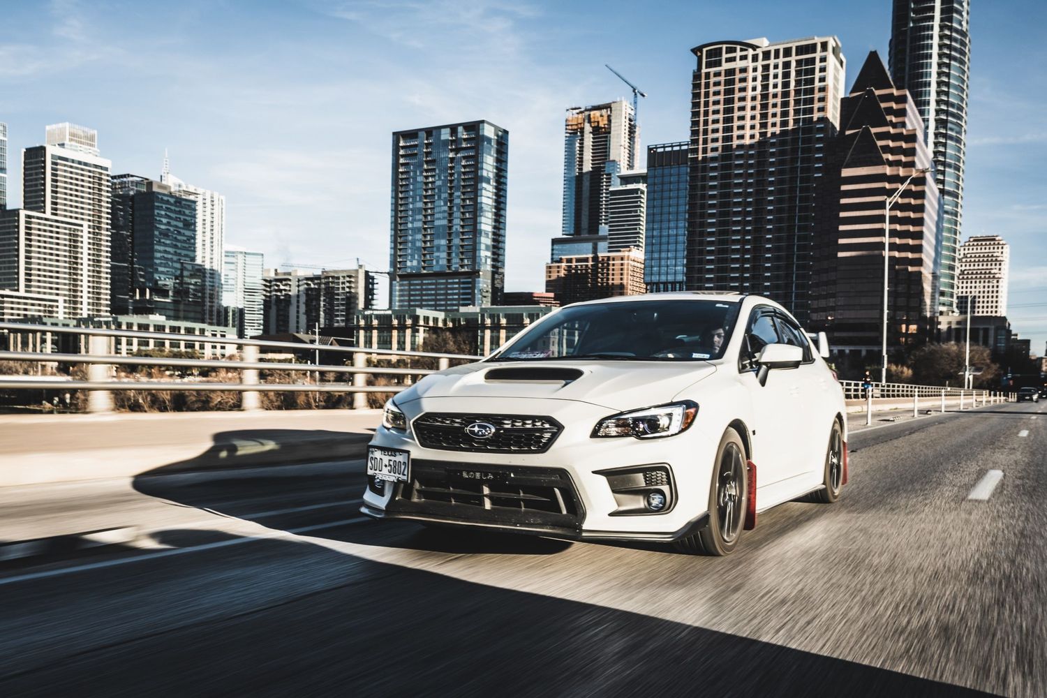 White Subaru WRX STI driving on road with modern city skyline and high-rise buildings in background.
