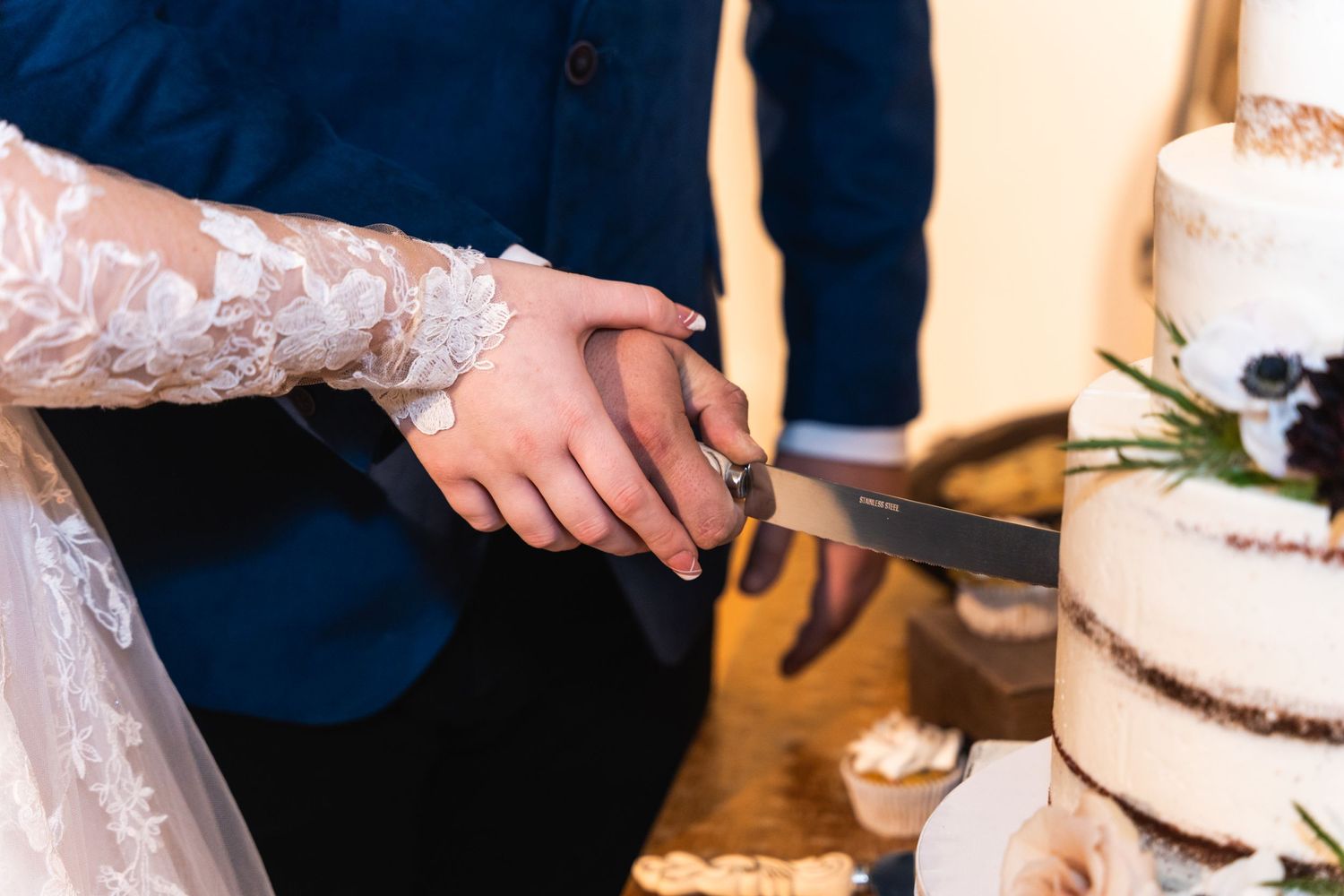 A close-up of hands cutting into a rustic white wedding cake decorated with lace details.
